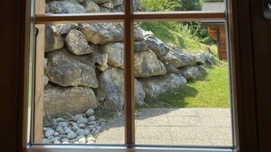 A view through a window with a stone wall and a wooden fence, overlooking a mountainous landscape and a building with a tiled roof. - Chalet - Calm, comfort and splendid view (Mayens de chamoson)