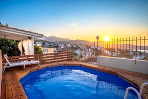 Une piscine sur le toit, dotée d’une terrasse en bois et de chaises longues blanches, offrant une vue sur le paysage urbain avec des montagnes en arrière-plan.
