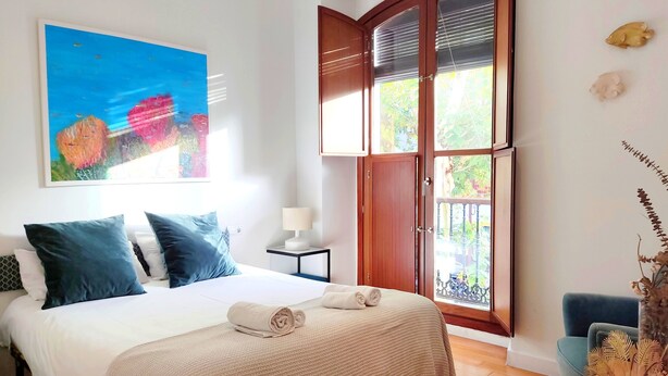 Um quarto com cama, mesa de cabeceira, janela com vista e um quadro na parede.