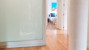 Um corredor com piso de madeira que leva a um quarto com cama e um quadro azul na parede.