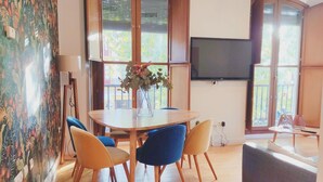 Uma área de refeições com uma mesa redonda, cadeiras azuis e amarelas, um vaso com flores e uma TV fixada na parede.