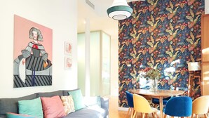 Uma sala de estar moderna com um sofá cinzento, almofadas coloridas, uma mesa de jantar com cadeiras e um vibrante papel de parede floral.