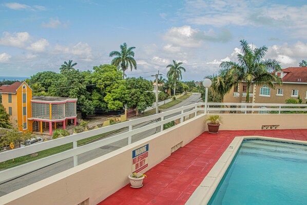 A swimming pool with a red deck, a lifeguard warning sign, and a view of surrounding buildings and trees.