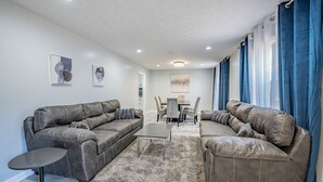 A modern living room with a sectional sofa, a small round table, and a dining area with a table and chairs. - Super Luxurious House (Atlanta)