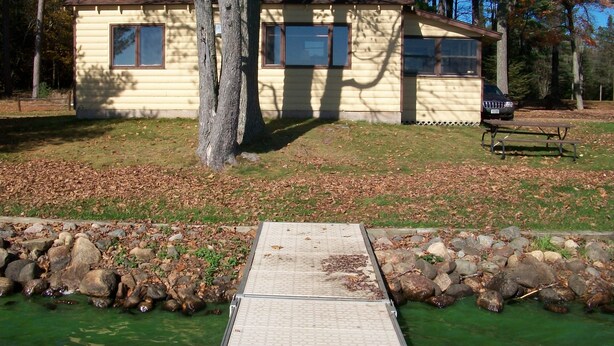 Een klein, gelijkvloers vakantiehuisje met een veranda, omgeven door bomen en een klein vijvertje met rotsen.