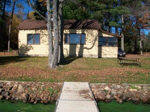 A small, single-story cabin with a porch, surrounded by trees and a small pond with rocks. - Cozy 2 Bedroom Cabin With Gas Fireplace (Saint Germain)