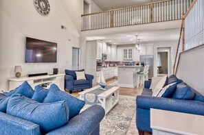A spacious living area with a high ceiling, a kitchen with white cabinets, and a dining area with white chairs.