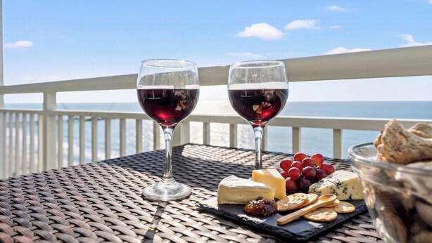Deux verres de vin rouge sur une table, avec vue sur la mer, accompagnés de fromage, de raisins et de crackers.