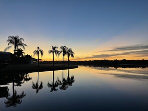 A calm lake at sunset with palm trees reflected in the water, a clear sky, and a distant shoreline.