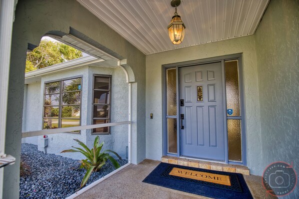 A house entrance with a blue door, a welcome mat, and a small garden area with pebbles and a plant.