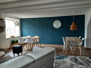 A modern living room with a sofa, a dining table with chairs, a clock on the wall, and a window with a view of trees.