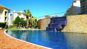 A swimming pool with a stone wall and a waterfall feature, surrounded by palm trees and a building with balconies. - Blue Sky Condo with Ocean&Pool view by Be Cherish (Albufeira)