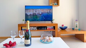A living room with a television displaying a coastal scene, a bottle of champagne, strawberries, and a bowl of seashells on a table. - Blue Sky Condo with Ocean&Pool view by Be Cherish (Albufeira)