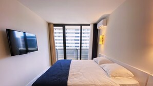 A hotel room with a bed, a TV mounted on the wall, a window with curtains, and a view of a building outside. - Orbi City/Two Rooms Apt./27 Floor/Sea - Mout. View (Batumi)