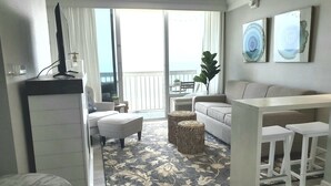 A modern living room with a sofa, ottoman, and coffee table, a balcony with a view, and artwork on the wall.
