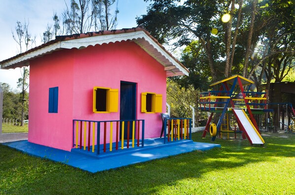 Un edificio rosa con puerta y ventanas azules, un porche azul y un parque infantil con tobogán y estructura para escalar.
