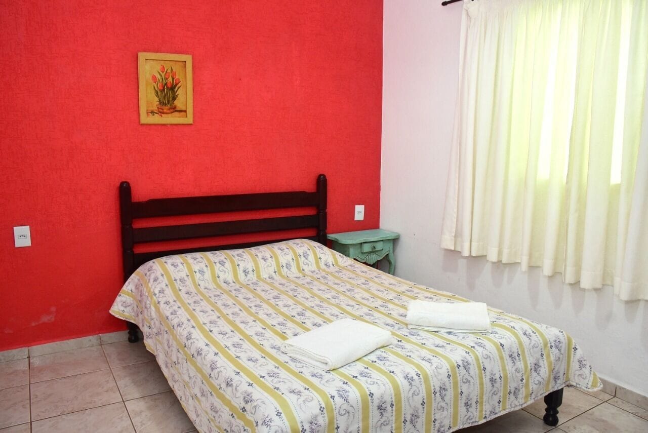 Uma cama com colcha listrada, uma mesa de cabeceira, uma parede vermelha e uma janela com cortinas.