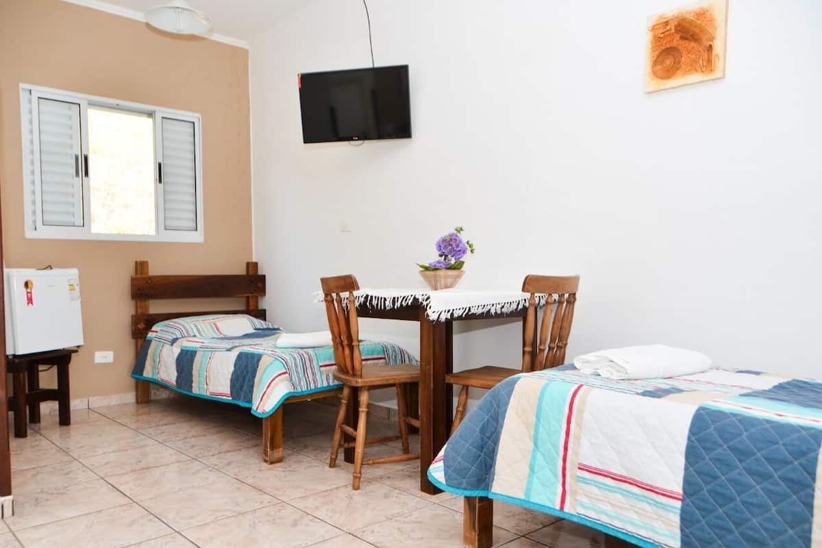 A room with two beds, a small table with chairs, a television on the wall, and a window with shutters.