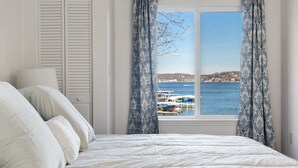 Ein Schlafzimmer mit Bett, Kissen, einem Fenster mit Vorhängen und Blick auf das Meer und Boote.