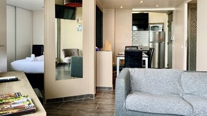 Et moderne hotelværelse med en grå sofa, et spiseområde med bord og stole, et køkken med køleskab og mikroovn, samt et badeværelse med toilet og håndvask.