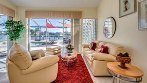 A spacious living room with a beige sofa, a coffee table, and a red rug. There is a large window with a view of a pool and outdoor furniture.