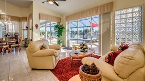 A spacious living room with a beige sofa, a coffee table, and a red rug. There is a dining area with wooden furniture and a large window offering a view of the outdoors.