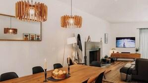 A modern living room with a wooden dining table, black chairs, a fireplace, a television, and contemporary lighting fixtures.