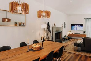 Ein modernes Wohnzimmer mit einem Holzesstisch, schwarzen Stühlen, einem Kamin, einem Fernseher und zeitgemäßen Leuchten.