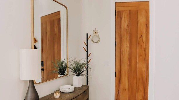 A wooden console table with a white lamp, a small plant, and a decorative item. A coat rack with a mirror and a coat is mounted on the wall. A wooden door is visible in the background.