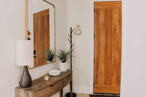 Une console en bois surmontée d’une lampe blanche, d’une petite plante et d’un objet décoratif. Un porte-manteau avec un miroir et un manteau est fixé au mur. On aperçoit une porte en bois à l’arrière-plan.