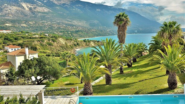 A pool area with palm trees and a view of a mountain and sea. - Seawave Villa (Καραβαδος)