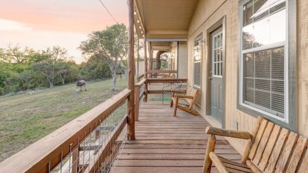 A wooden porch with a railing, a bench, and a view of a grassy area with trees and a barbecue grill. - Gone Fishin` Cabin at Lightning Bug Springs (Uvalde)