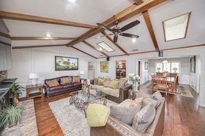 A spacious living room with a high ceiling, wooden beams, a ceiling fan, and a dining area with a wooden table and chairs.