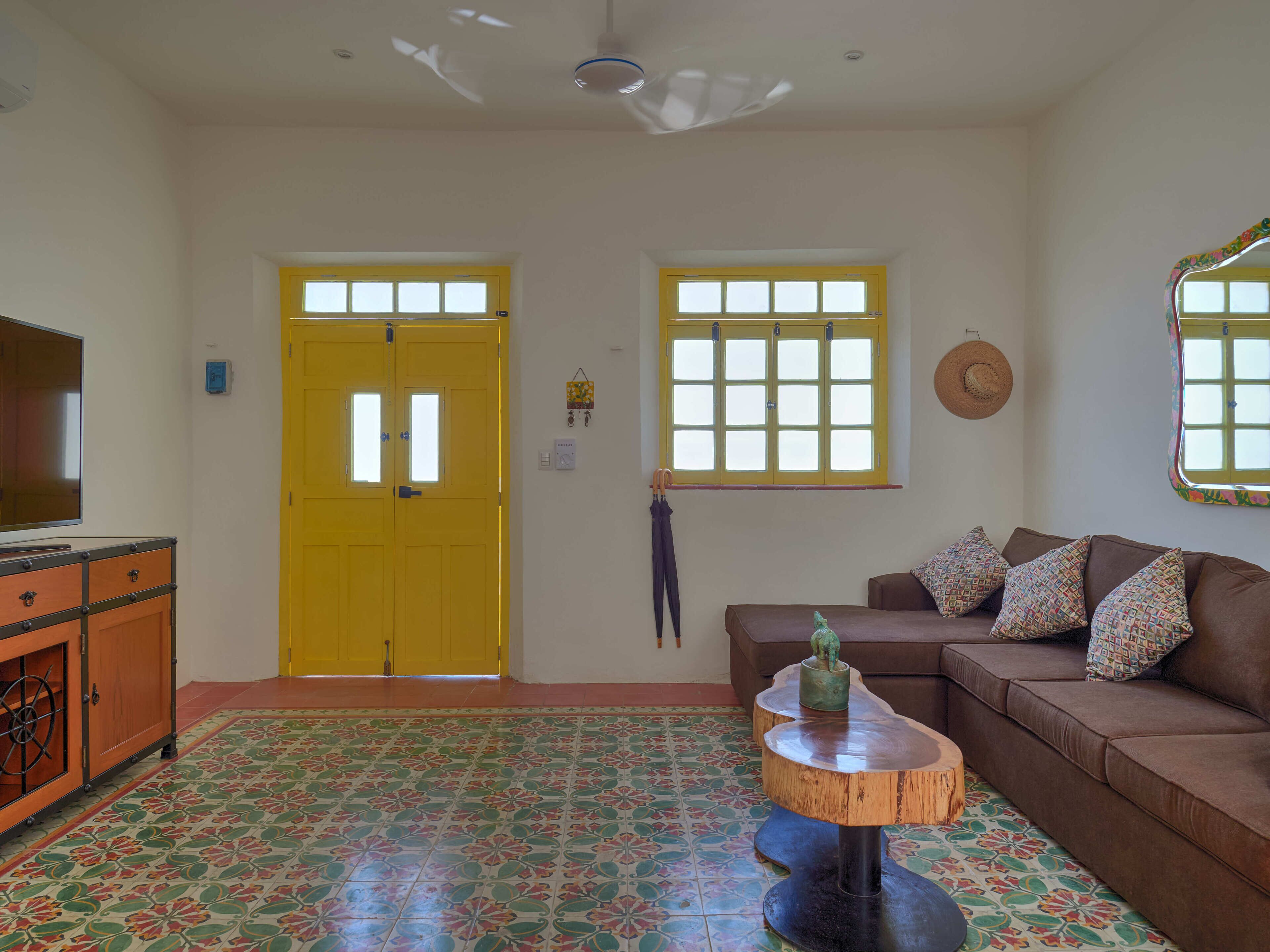A living room with a brown sofa, a wooden coffee table, a TV on a cabinet, a yellow door, and a window with a decorative frame.