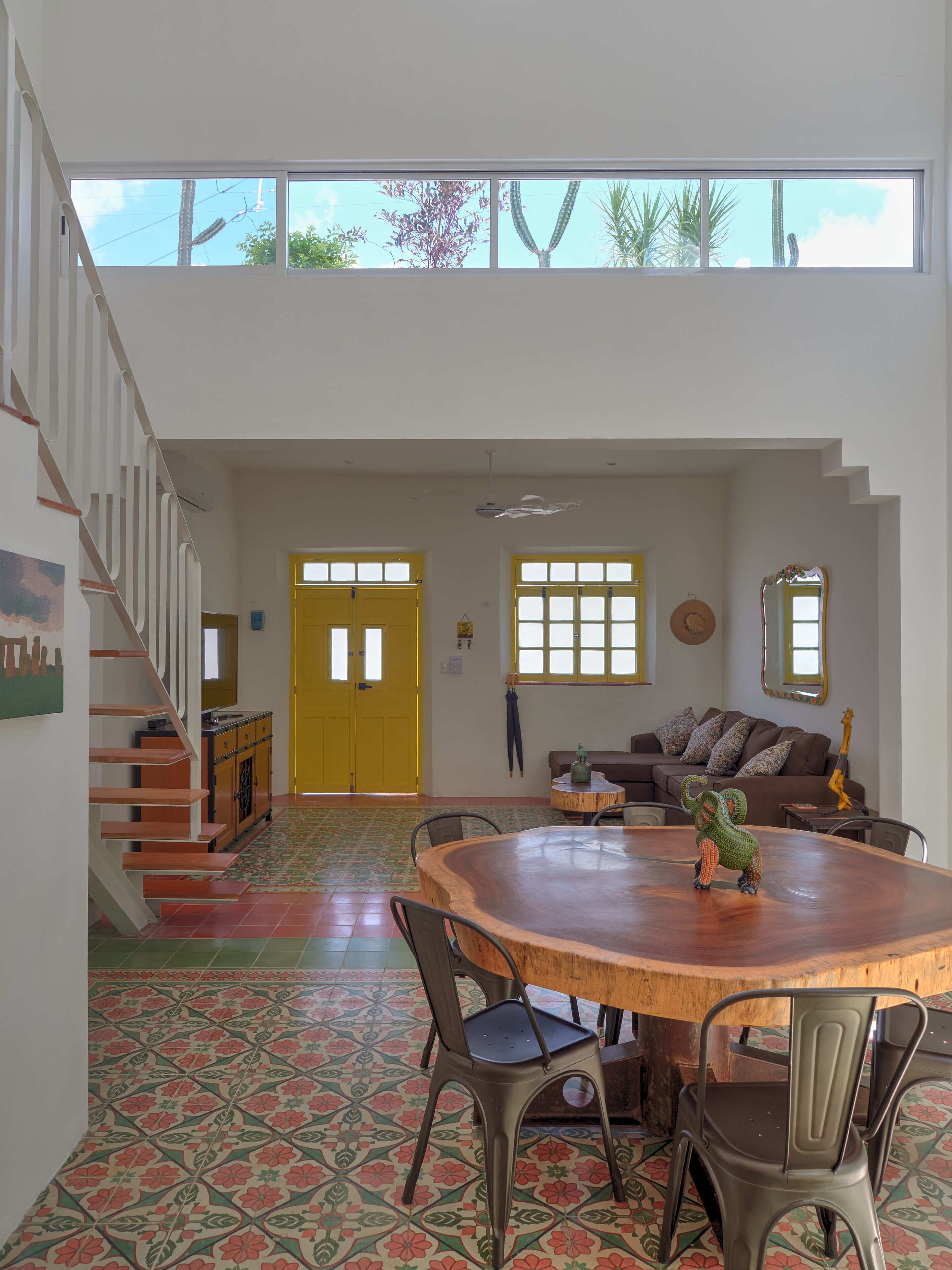 A spacious living area with a high ceiling, a wooden dining table surrounded by chairs, a staircase with a railing, and a yellow door.