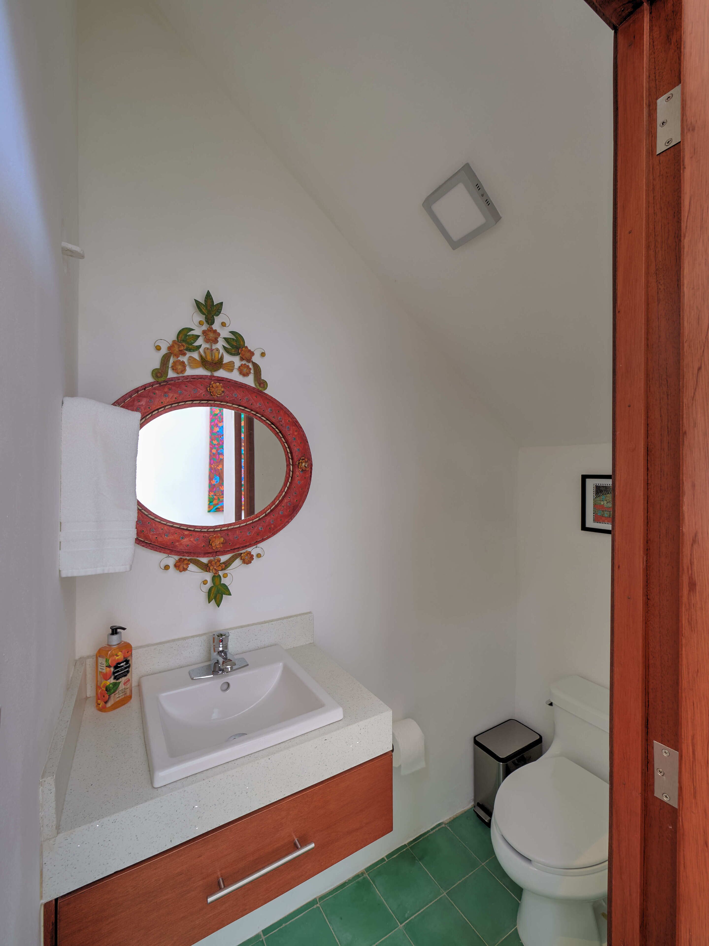 A bathroom with a white sink, a mirror with a floral frame, a toilet, and a trash bin.