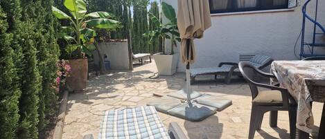 An outdoor patio area with a stone floor, a lounge chair, a table with a patterned tablecloth, a covered umbrella, and potted plants.