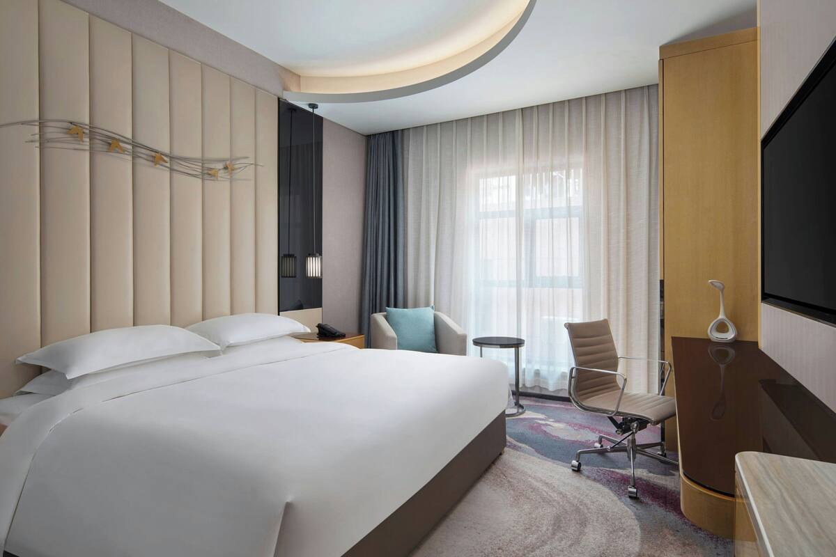 Quarto de hotel moderno com uma cama grande, uma escrivaninha com cadeira, uma mesinha e uma televisão.