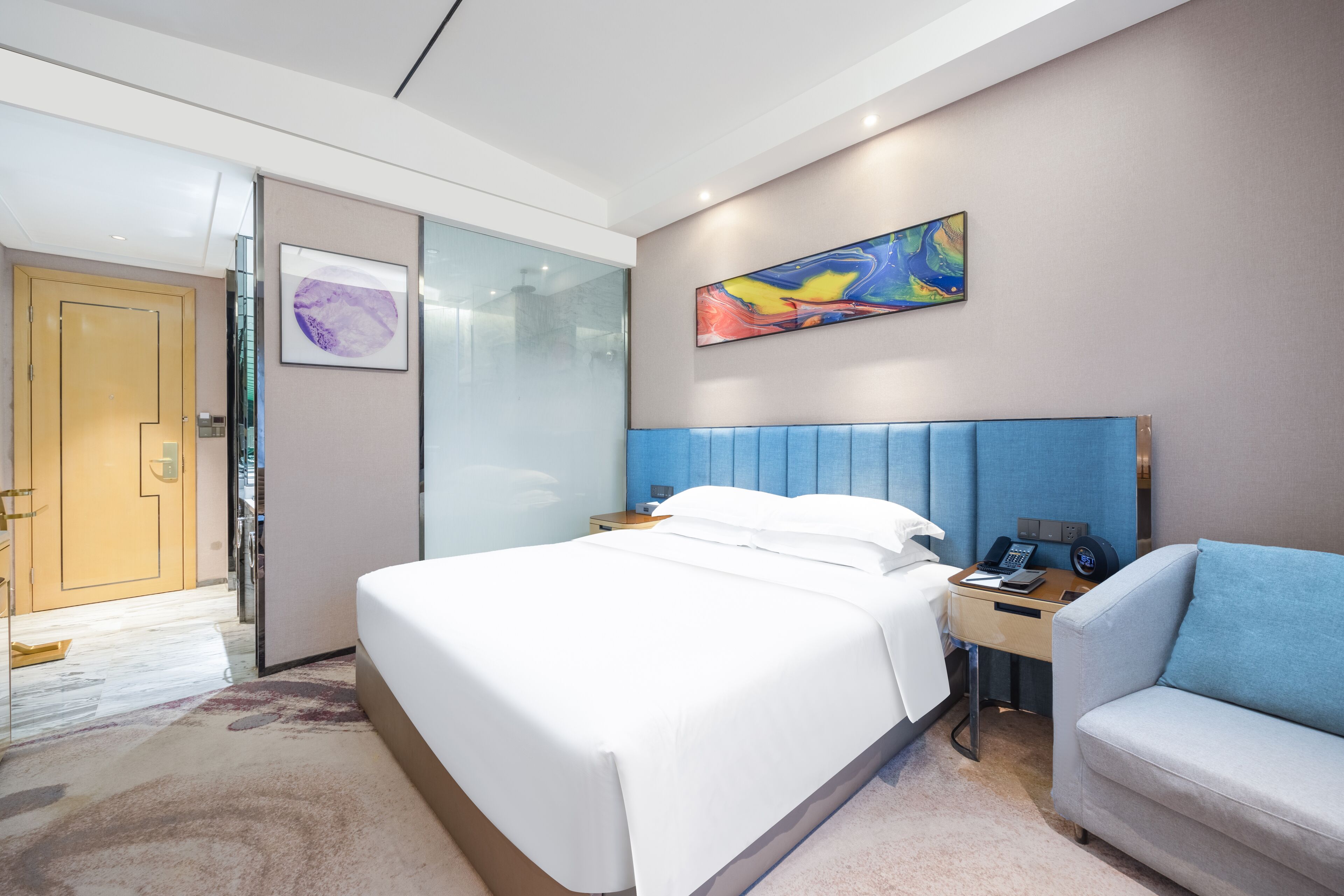 A modern hotel room with a large bed, a desk with a chair, a glass door, and a colorful abstract painting on the wall.