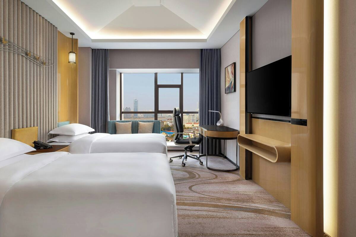 Quarto de hotel com duas camas, uma escrivaninha, uma cadeira, televisão e uma grande janela com vista para a cidade.
