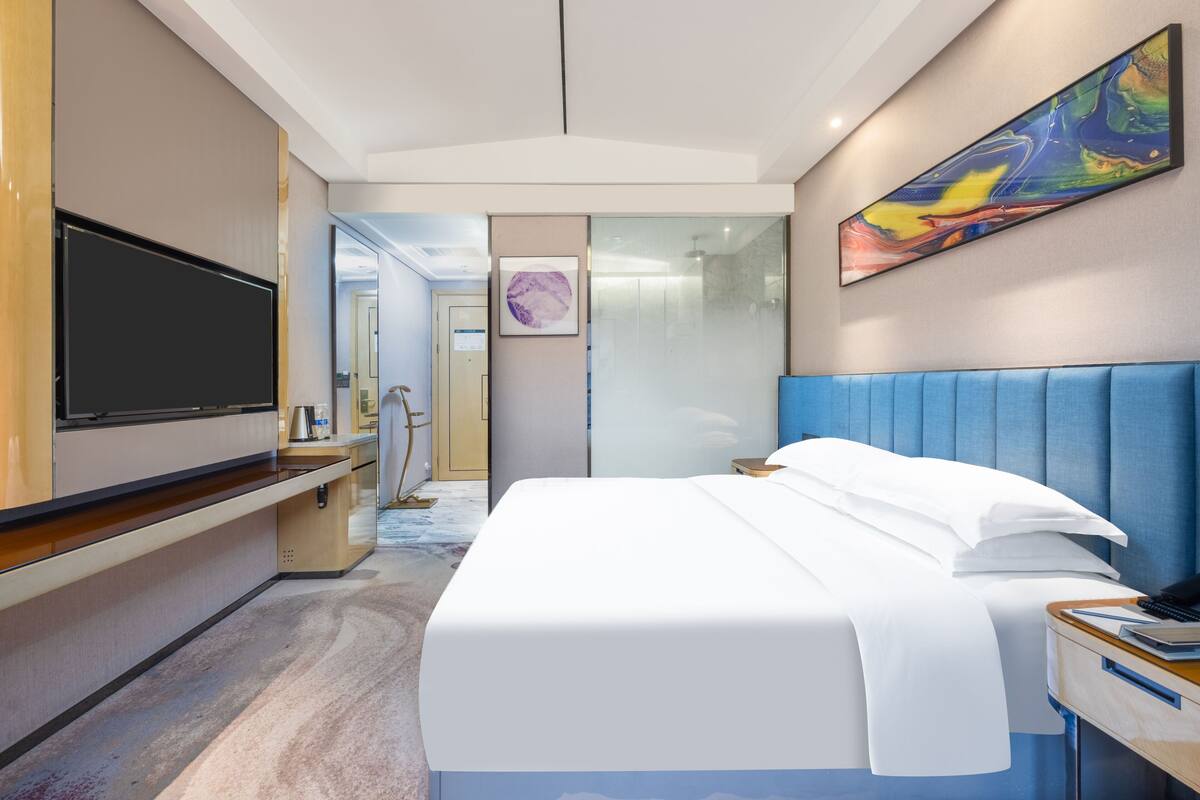 Quarto de hotel moderno com uma cama grande, televisão de tela plana, escrivaninha com cadeira e banheiro visível por uma porta de vidro.