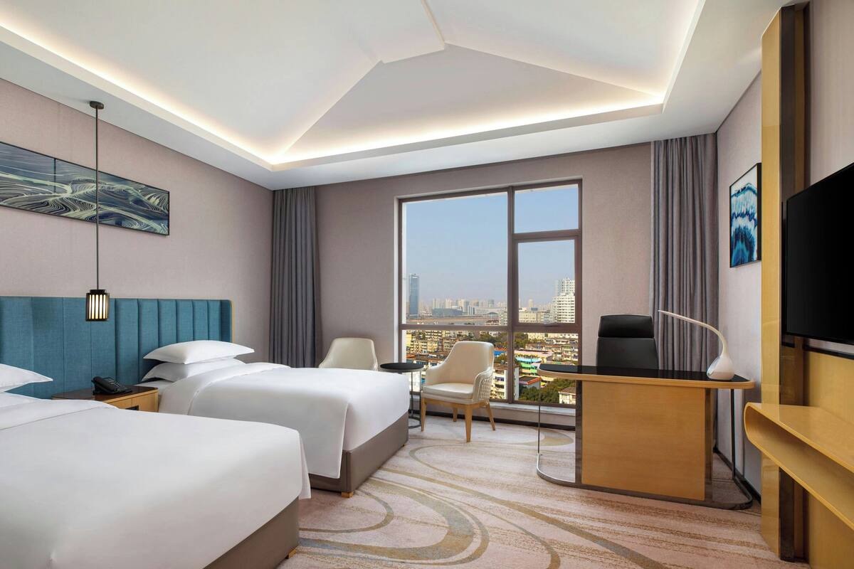 Quarto de hotel com duas camas, uma escrivaninha, uma cadeira, televisão e uma janela grande com vista para a cidade.