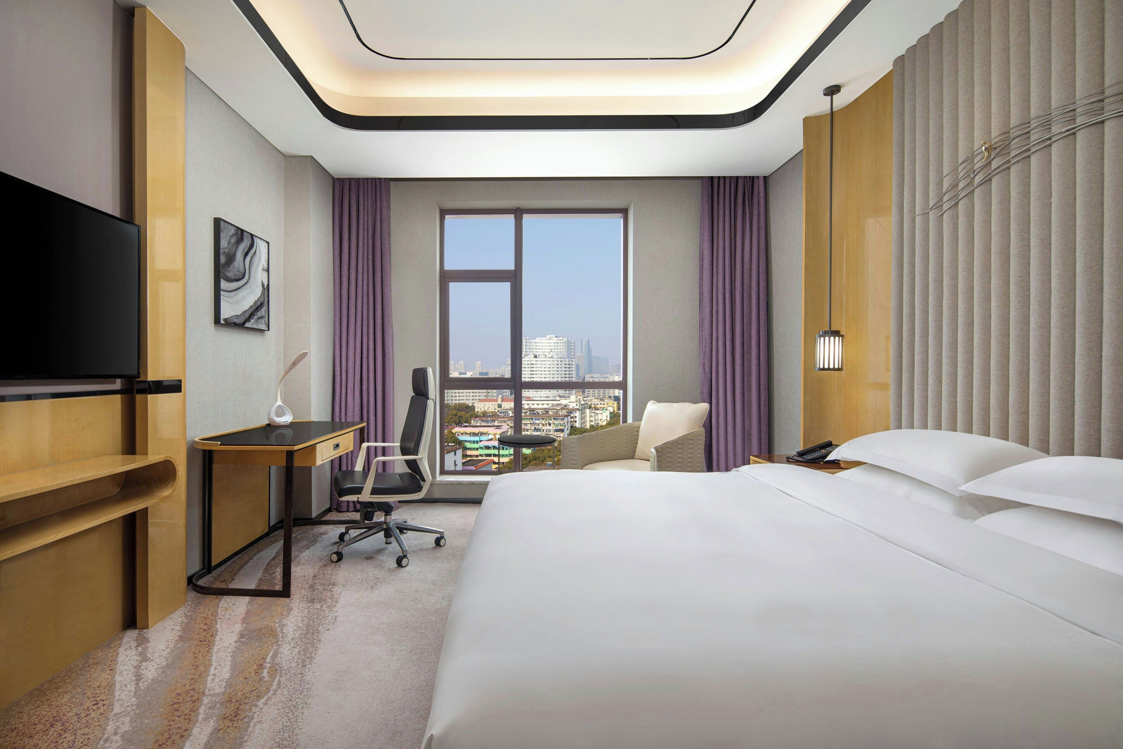 Habitación de hotel con una cama grande, un escritorio con silla, televisor y vistas de la ciudad a través de un ventanal amplio.