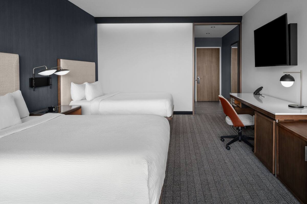 A hotel room with two beds, a desk, a chair, a TV, and a lamp.