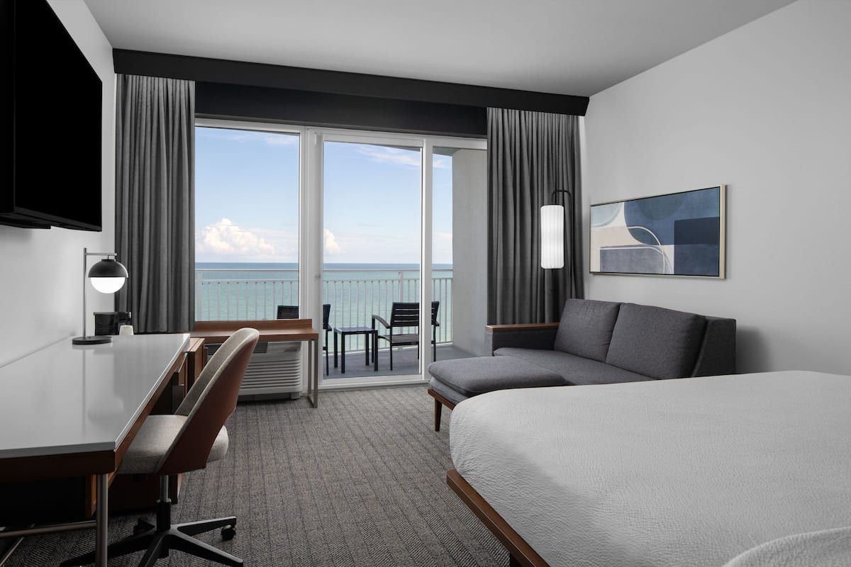 A hotel room with a bed, a desk, a chair, a TV, and a balcony with a view of the sea.