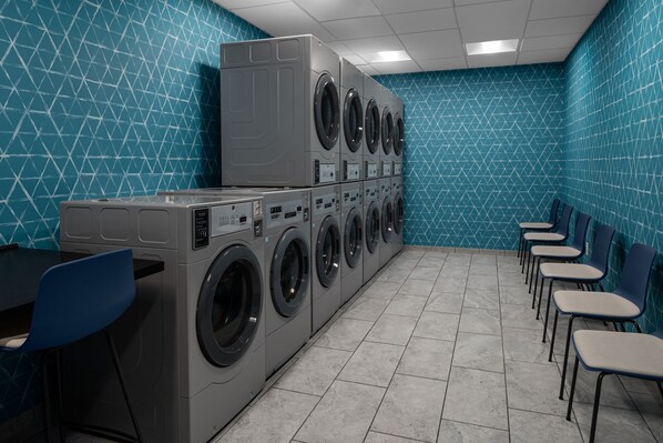 Une buanderie équipée de machines à laver et de sèche-linge superposés, un espace détente avec des chaises et un sol carrelé.