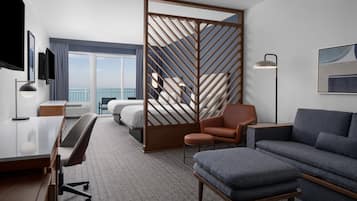 A modern hotel room with a bed, a desk, a sofa, and a large window offering a view of the sea.
