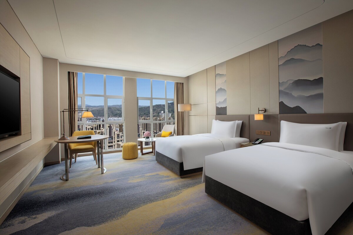 A hotel room with two beds, a TV, a desk with a chair, and a view of the city through large windows.