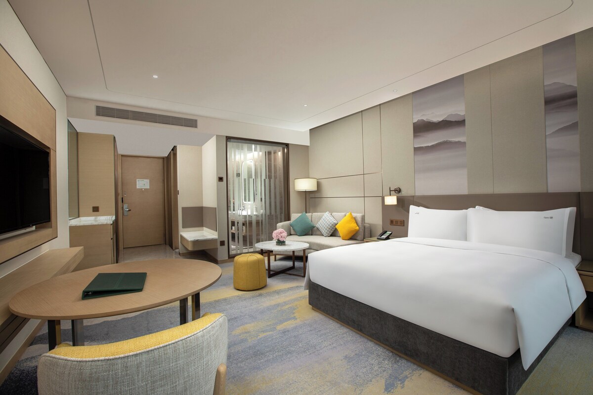 A hotel room with a bed, a desk, a chair, a TV, and a wall with a painting.