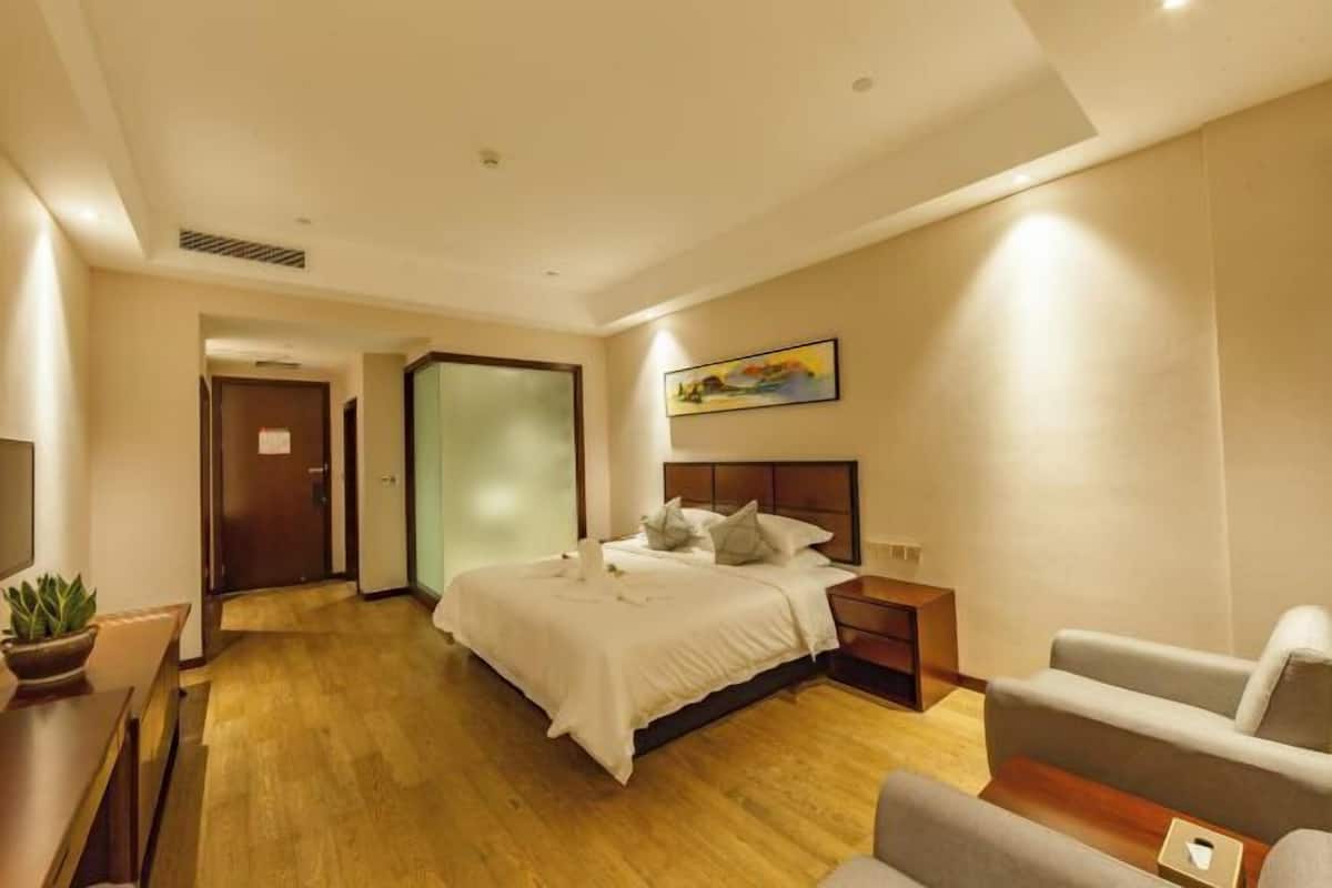 A hotel room with a bed, a bedside table, a chair, a desk, and a potted plant.